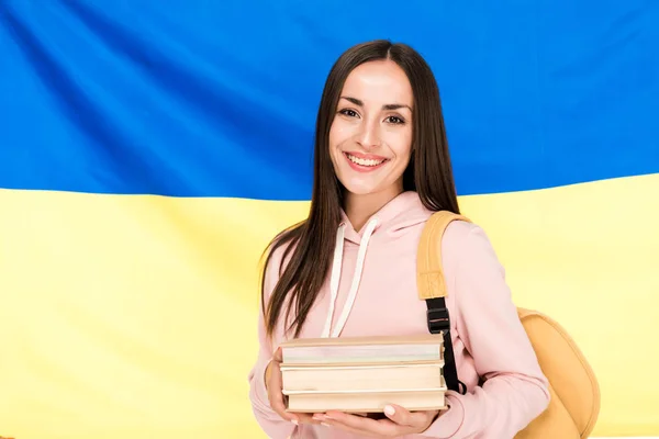 Lächelnde brünette junge Frau mit Rucksack und Büchern auf ukrainischem Fahnenhintergrund — Stockfoto