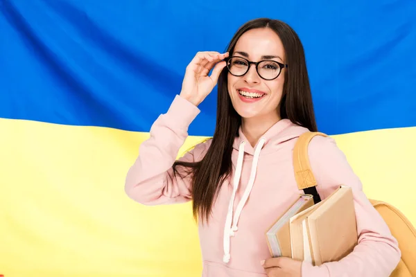 Jeune femme brune souriante avec sac à dos et livres touchant des lunettes sur fond drapeau ukrainien — Photo de stock
