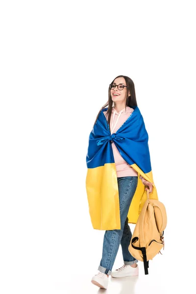 Piena vista lunghezza di felice ragazza in occhiali camminare con lo zaino e la bandiera dell'Ucraina sulle spalle isolate su bianco — Foto stock