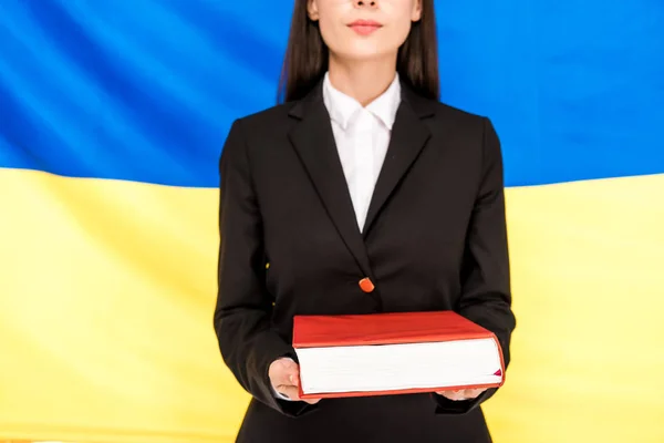 Vue recadrée de l'avocat en costume noir tenant livre sur fond drapeau ukrainien — Photo de stock