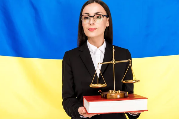 Abogado en traje negro y gafas que sostienen escalas de justicia en el libro sobre fondo de la bandera de Ucrania - foto de stock