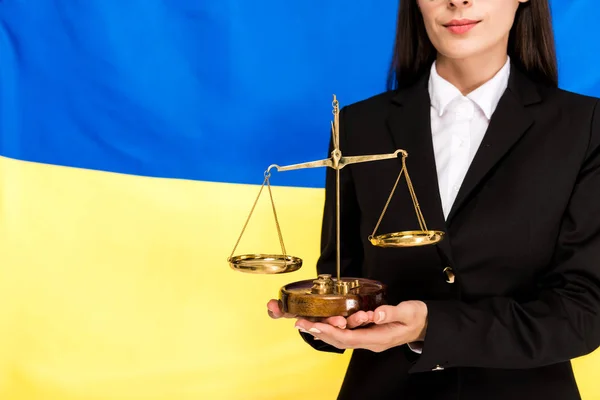 Vista cortada de advogado em terno preto segurando escalas de justiça no fundo bandeira ucraniana — Fotografia de Stock