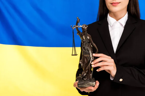 Vue recadrée de l'avocat en costume noir tenant statuette Themis sur fond drapeau ukrainien — Photo de stock