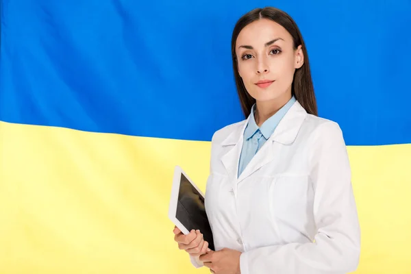 Врач в белом халате держит планшет на фоне украинского флага — стоковое фото
