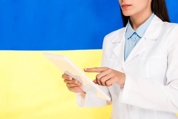 Vista recortada del médico de bata blanca con tableta digital sobre fondo de la bandera de Ucrania - foto de stock