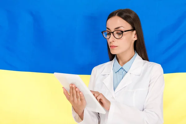 Лікар у білому пальто та окуляри використовуючи цифровий планшет на фоні українського прапора — стокове фото