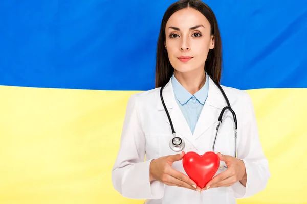 Врач в белом халате со стетоскопом с красным сердцем на фоне украинского флага — стоковое фото
