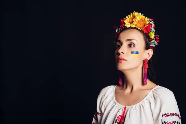 Brunette young woman in national Ukrainian costume and floral wreath looking away isolated on black — Stock Photo