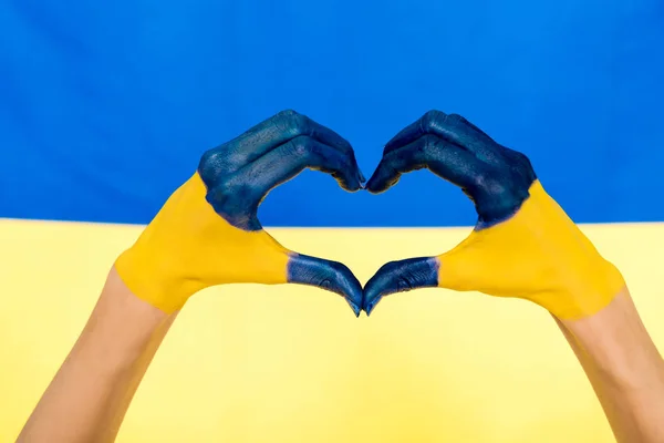 Vista recortada de las manos pintadas mostrando el signo del corazón en el fondo de la bandera de Ucrania - foto de stock