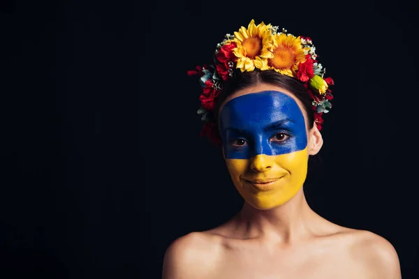 Naked young woman in floral wreath with painted Ukrainian flag on skin smiling isolated on black — Stock Photo