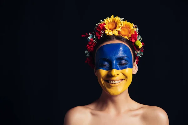 Naked young woman in floral wreath with painted Ukrainian flag on skin smiling with closed eyes isolated on black — Stock Photo