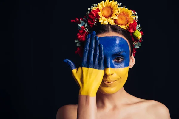 Giovane donna nuda sorridente in ghirlanda floreale con bandiera ucraina dipinta su pelle con mano sul viso isolato su nero — Foto stock