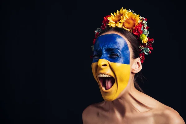 Naked young woman in floral wreath with painted Ukrainian flag on skin screaming isolated on black — Stock Photo