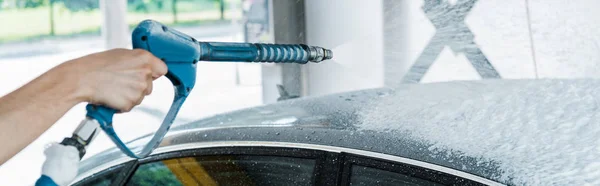 Panoramic shot of car washer holding pressure washer with foam near car — Stock Photo