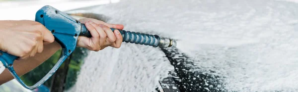 Panoramic shot of car cleaner holding pressure washer with foam near car — Stock Photo