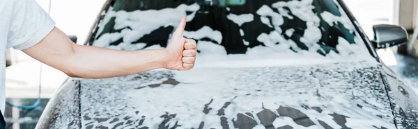 Panoramic shot of car cleaner showing thumb up near car with foam — Stock Photo