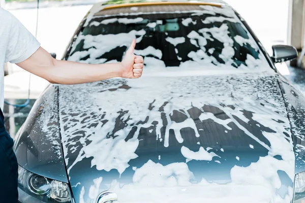Cropped view of car cleaner showing thumb up near car with foam — Stock Photo