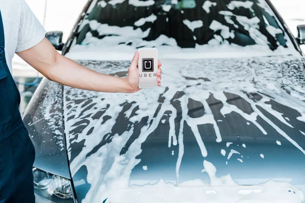 KYIV, UCRAINA - 20 GIUGNO 2019: vista ritagliata dell'uomo che tiene smartphone con app uber sullo schermo vicino all'auto con schiuma — Foto stock