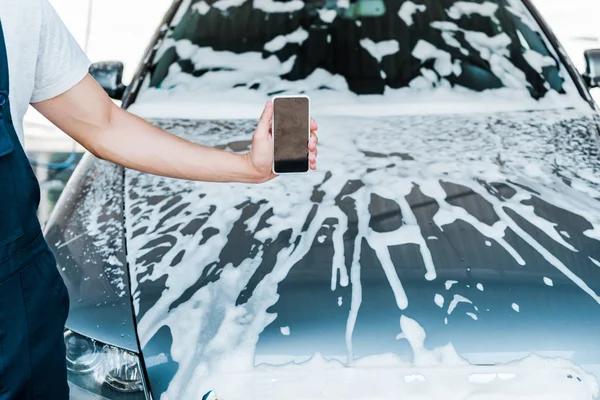 Обрезанный вид человека, держащего смартфон с пустым экраном рядом с машиной с пеной — стоковое фото