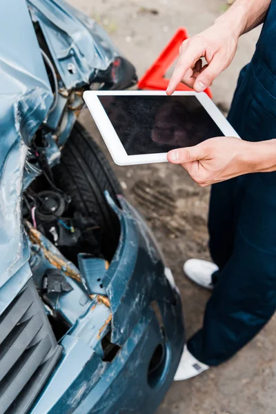 Обрезанный вид человека, указывающего пальцем на цифровой планшет с пустым экраном рядом с поврежденным автомобилем — стоковое фото