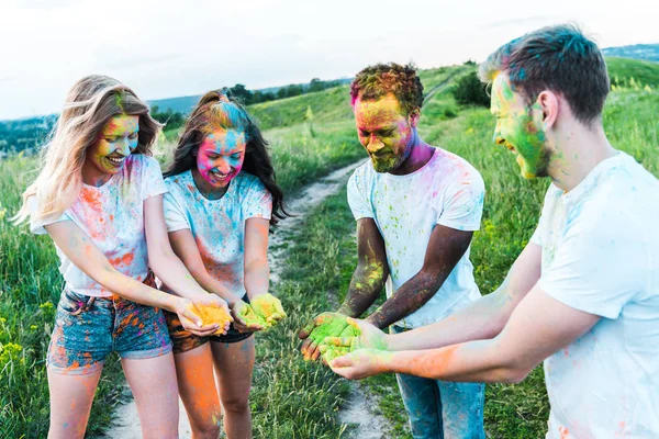 Felices amigos multiculturales sosteniendo coloridas pinturas holi en las manos - foto de stock