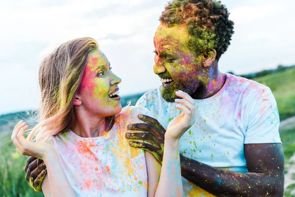 Hombre afroamericano feliz tocando hombros de mujer joven sorprendida con pinturas holi en la cara - foto de stock