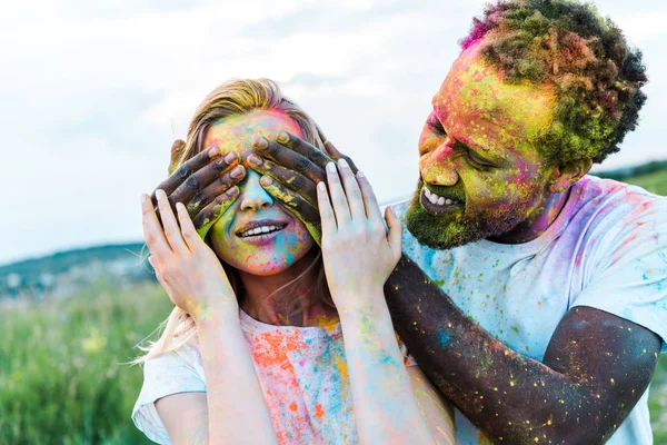 Homem americano africano feliz cobrindo olhos de mulher jovem com tintas holi no rosto — Fotografia de Stock