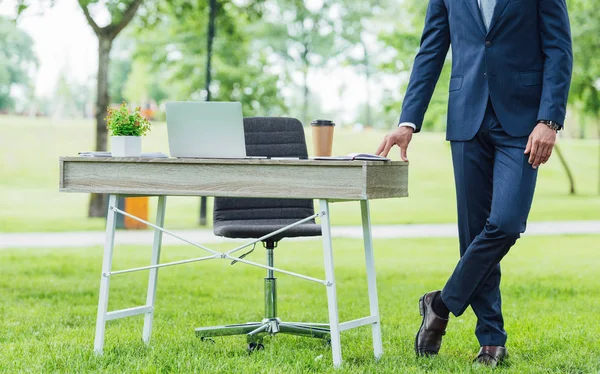 Vista recortada del joven empresario de pie con las piernas cruzadas cerca de la mesa en el parque — Stock Photo