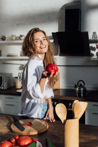 Laughing young woman in white shirt holding tomato in kitchen — Stock Photo