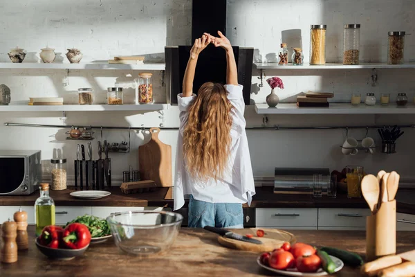 Vista posteriore di giovane donna in camicia bianca in piedi con le mani in alto in cucina — Foto stock