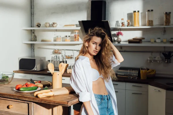 Sexy joven mujer en blanco sujetador de pie cerca de la mesa y mirando a la cámara en la cocina - foto de stock