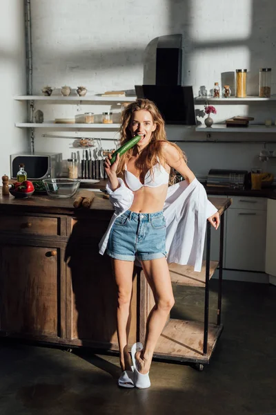 Ansicht des sexy Mädchens, das in der Nähe des Tisches steht und Gurken in der Küche isst — Stockfoto
