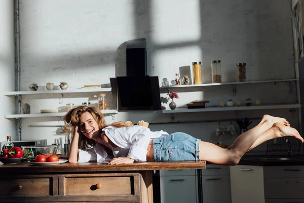 Sonriente sexy descalza joven mujer acostada en la mesa y mirando a la cámara en la cocina - foto de stock