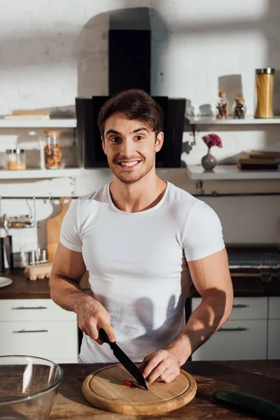 Hombre musculoso sonriente en camiseta blanca cortando verduras en la cocina - foto de stock