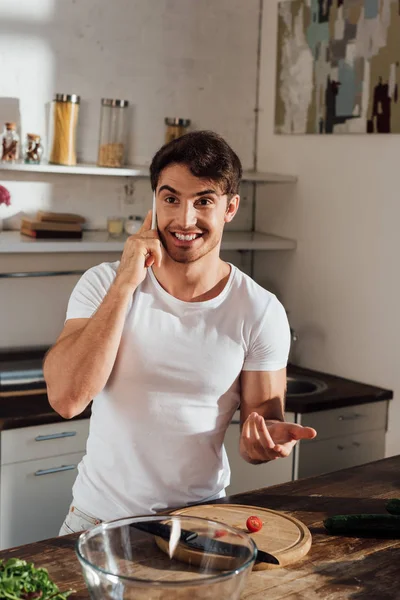 Homme souriant parler sur smartphone tout en coupant des tomates cerises dans la cuisine — Photo de stock