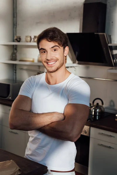 Homme musclé en t-shirt blanc debout avec les bras croisés et souriant dans la cuisine — Photo de stock