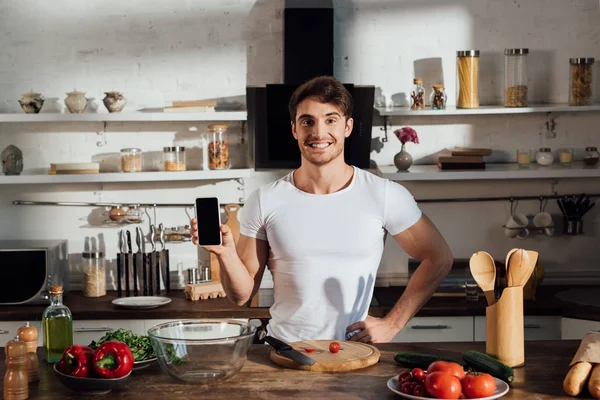 Vista frontal del hombre musculoso sonriente en camiseta blanca que muestra el teléfono inteligente con pantalla en blanco mientras cocina en la cocina - foto de stock