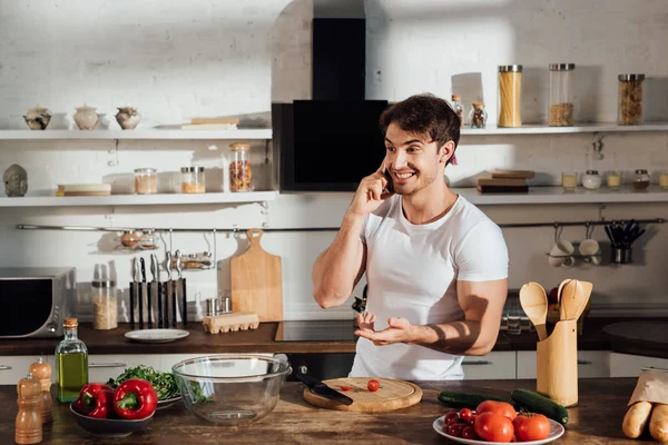 Homem muscular sorridente em t-shirt branca falando no smartphone enquanto cozinha na cozinha — Fotografia de Stock