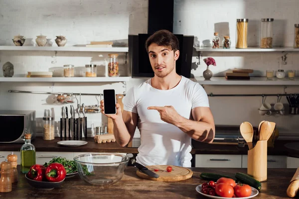 Homme musclé en t-shirt blanc pointant du doigt le smartphone avec écran blanc pendant la cuisson dans la cuisine — Photo de stock