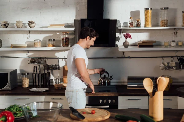 Sorridente muscular homem em branco t-shirt colocando chaleira no forno na cozinha — Fotografia de Stock