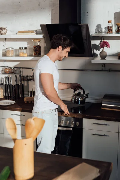 Улыбающийся мускулистый мужчина в белой футболке кладет чайник на духовку на кухне — стоковое фото
