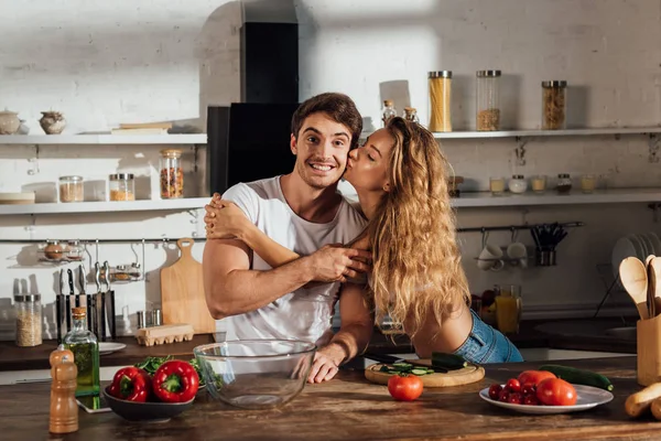 Sexy girl kissing smiling boyfriend near table in kitchen — Stock Photo