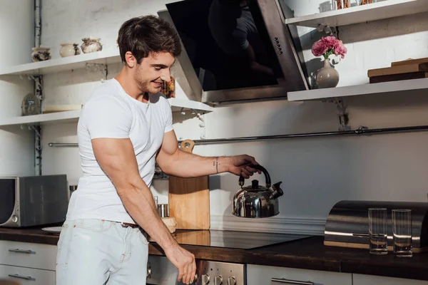 Hombre musculoso sonriente en camiseta blanca poner hervidor de agua en el horno en la cocina - foto de stock
