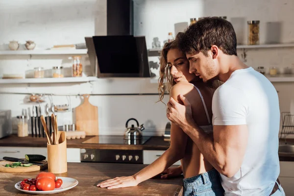 Musculoso hombre abrazando sexy chica en blanco sujetador en cocina - foto de stock