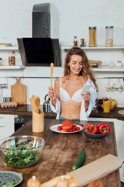 Усміхнена жінка в білому бюстгальтері використовує смартфон під час приготування їжі на кухні — стокове фото