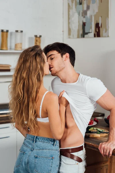 Sexy girl in white bra undressing and kissing boyfriend in kitchen — Stock Photo