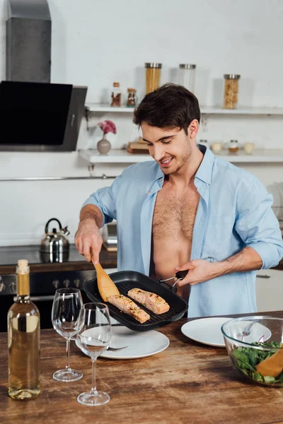 Sexy smiling man holding frying pan with fish in kitchen — Stock Photo