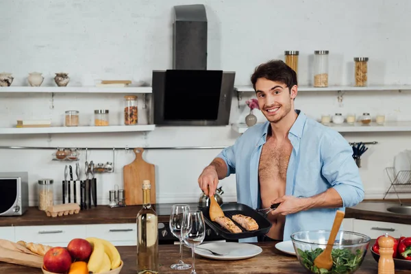 Sexy smiling man holding frying pan with fish in kitchen — Stock Photo