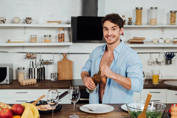 Lächeln sexy Mann öffnet Flasche Wein und blickt in die Kamera in der Küche — Stockfoto