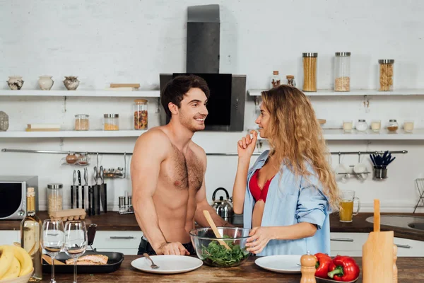 Sexy sorridente coppia in piedi vicino al tavolo con il cibo e guardando l'un l'altro in cucina — Foto stock